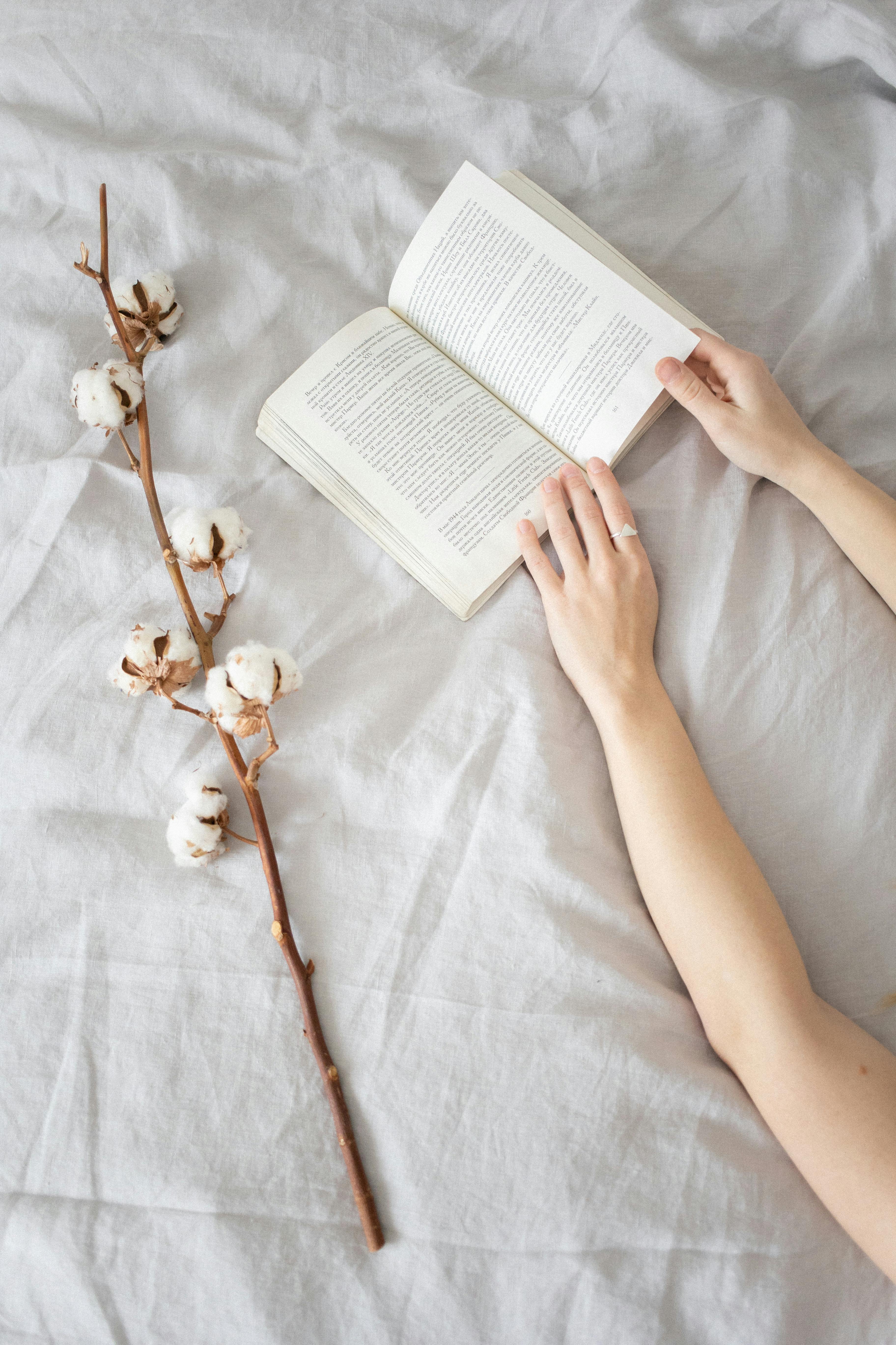 Hands flipping pages of a book next to cotton branch on a soft bed, creating a peaceful scene.