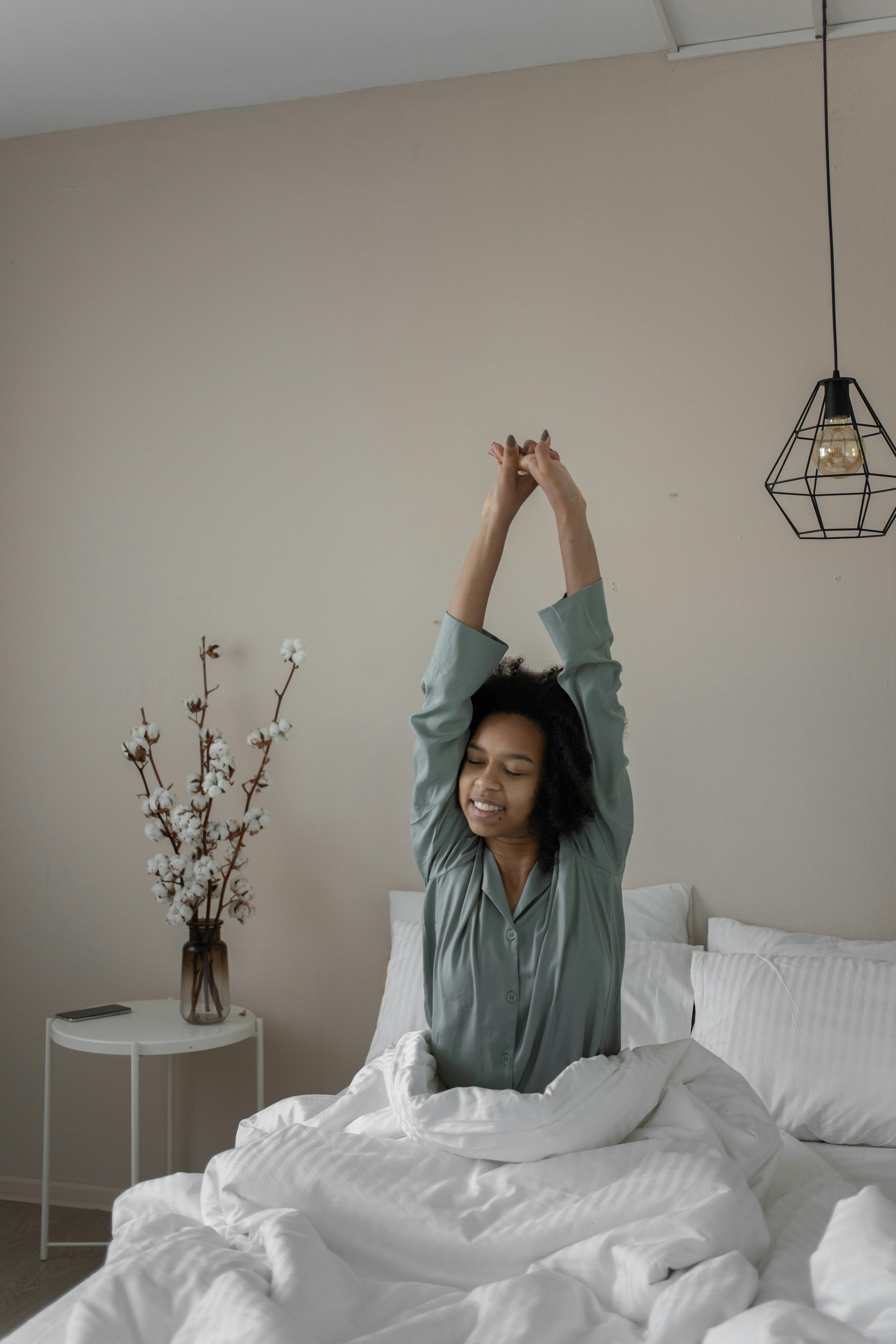 A young woman stretches with a smile, sitting in a cozy bedroom in the morning light.