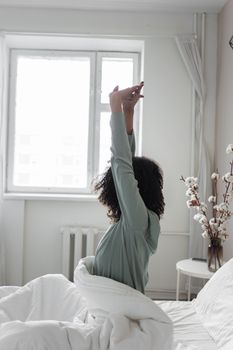 A woman with curly hair stretches in a cozy minimalist bedroom, embracing morning light.