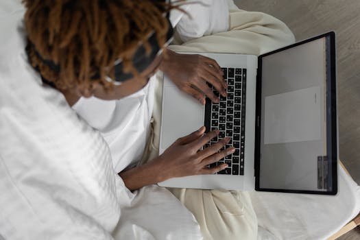 Person typing on a laptop while relaxing in bed. Minimalist, cozy setting with a focus on work-from-home lifestyle.