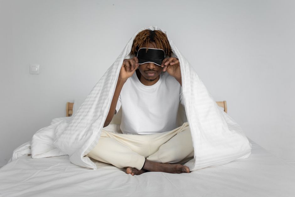 African American man sitting on bed with a sleep mask, wrapped in a comforter for relaxation. African American man sitting on bed with a sleep mask, wrapped in a comforter for relaxation.