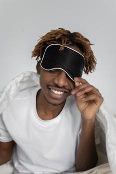 Young black man with dreadlocks smiling and adjusting his sleep mask in a cozy indoor setting.