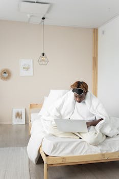 Young man with dreadlocks browsing a laptop while sitting on a bed in a modern bedroom.