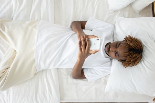 A young man with dreadlocks lying on bed, using a smartphone, wearing a white shirt.