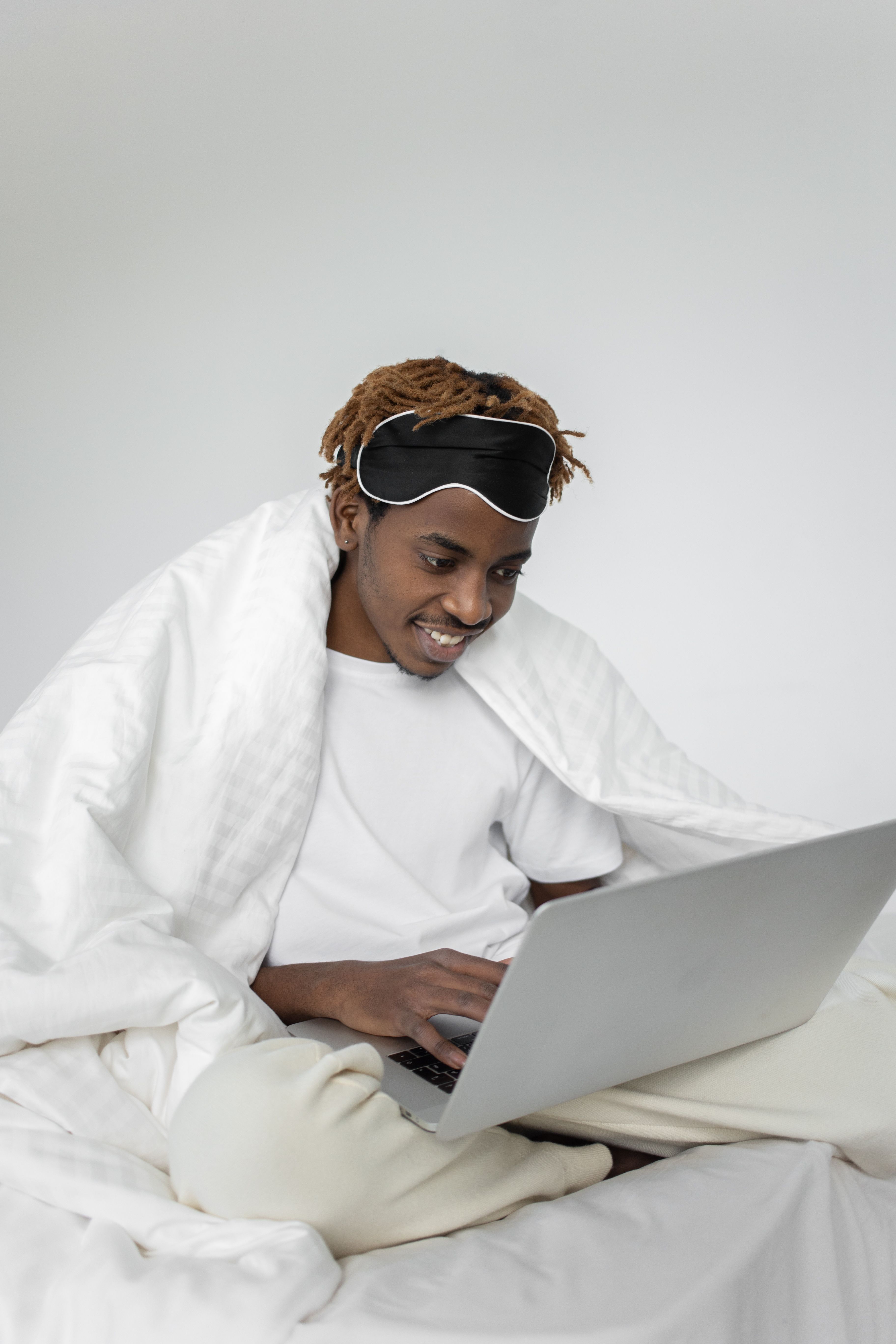 Smiling man with sleep mask working on a laptop in bed.