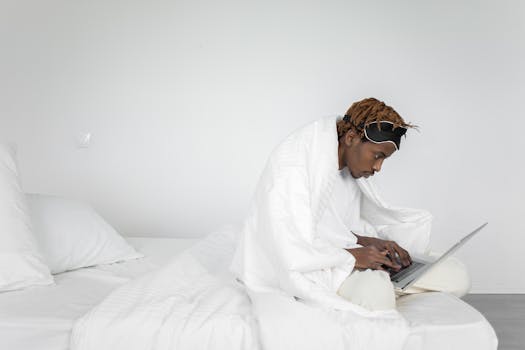 A man sits in bed with a laptop, working while wearing a sleep mask. Minimalist bedroom setting.