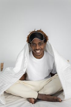 A cheerful man with a sleep mask sits on a bed, wrapped in a white blanket.