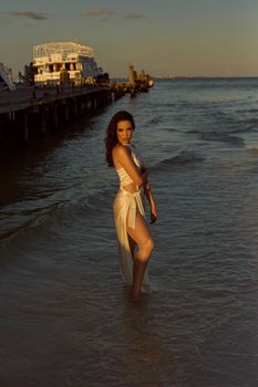 A woman stands gracefully on a beach at sunset, showcasing fashion and serenity by the sea.
