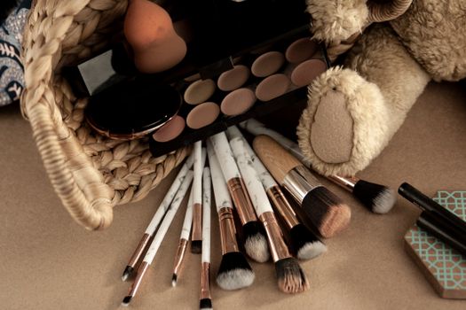 From above of various cosmetic products and makeup brushes in wicker basket placed near toy on beige table