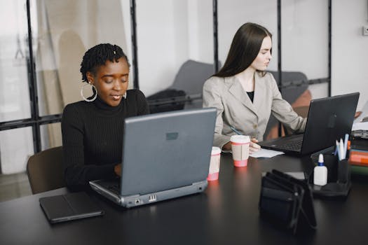 Two women working together on laptops in a modern office environment, showcasing teamwork and diversity.