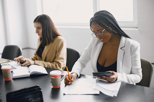 Two women working together in an office, taking notes and using a laptop.