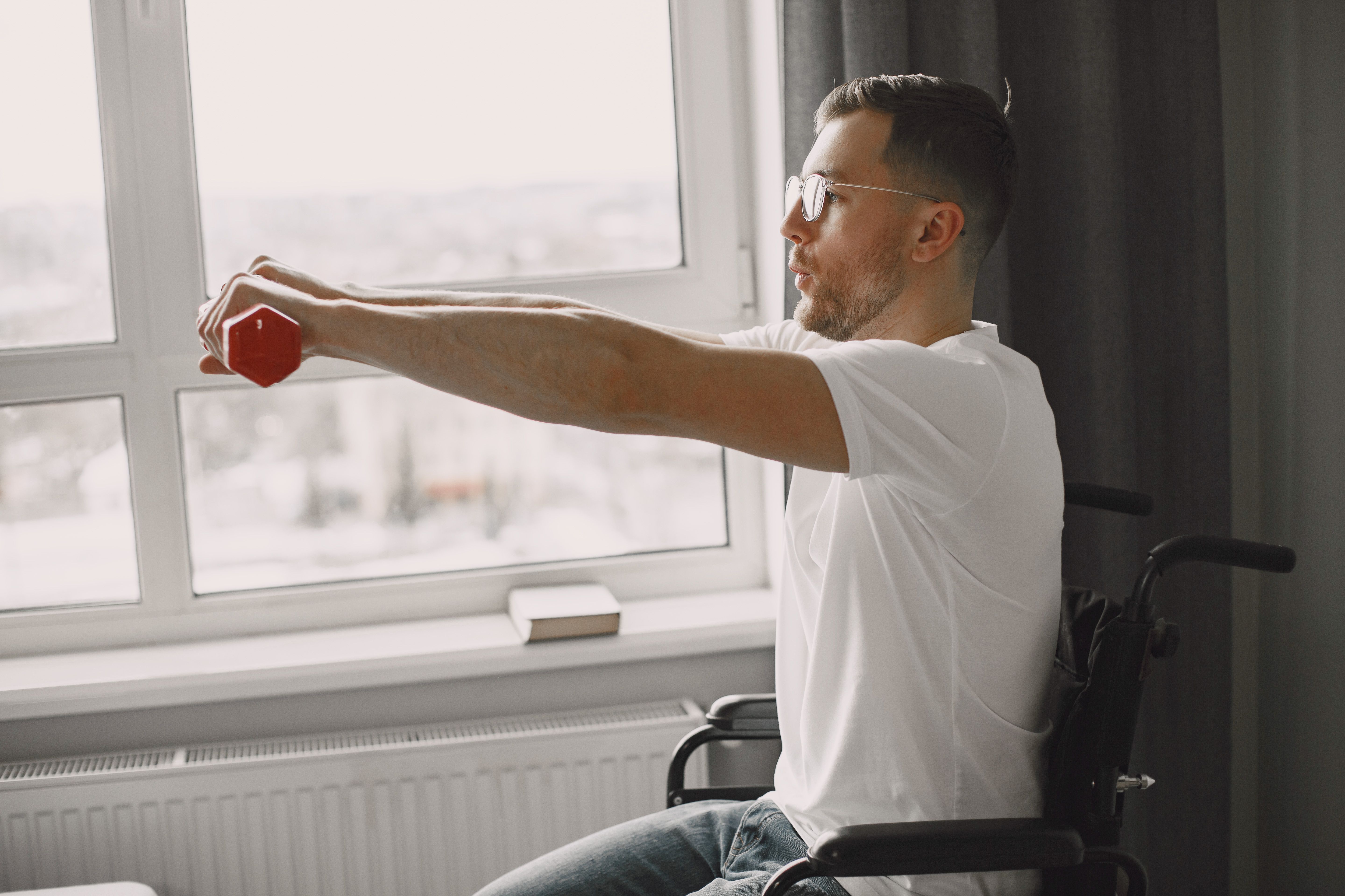 A man in a wheelchair exercising with a dumbbell by a bright window, promoting inclusivity and health.
