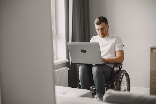 Adult man seated in a wheelchair and typing on a laptop, in a bright indoor setting.