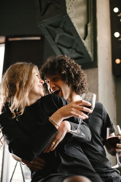 Free Happy couple laughing and holding wine glasses during a fun indoor moment. Stock Photo