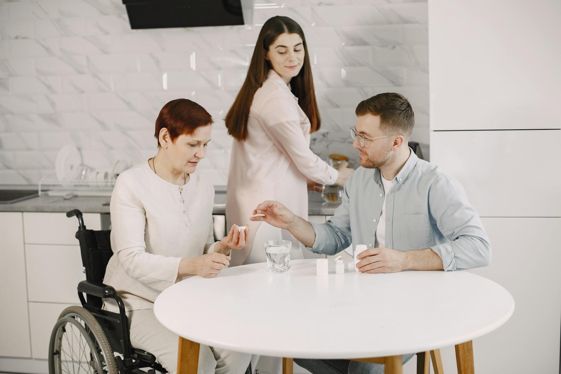 Elderly woman in wheelchair receiving help with medication from caregiver in modern kitchen.