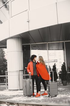 Couple with suitcases standing outside a modern building, embracing in winter attire.