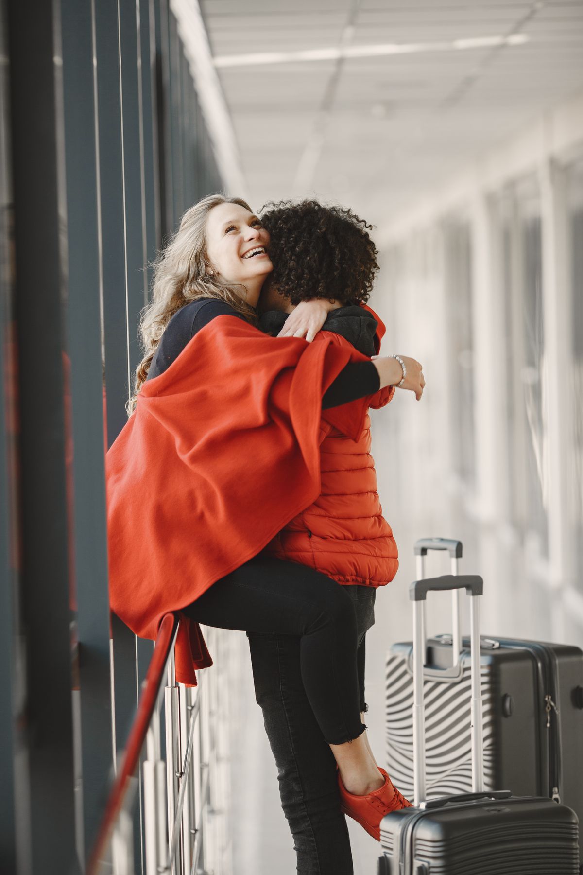 Two women sharing a joyful hug reunion in an airport corridor, with suitcases beside them