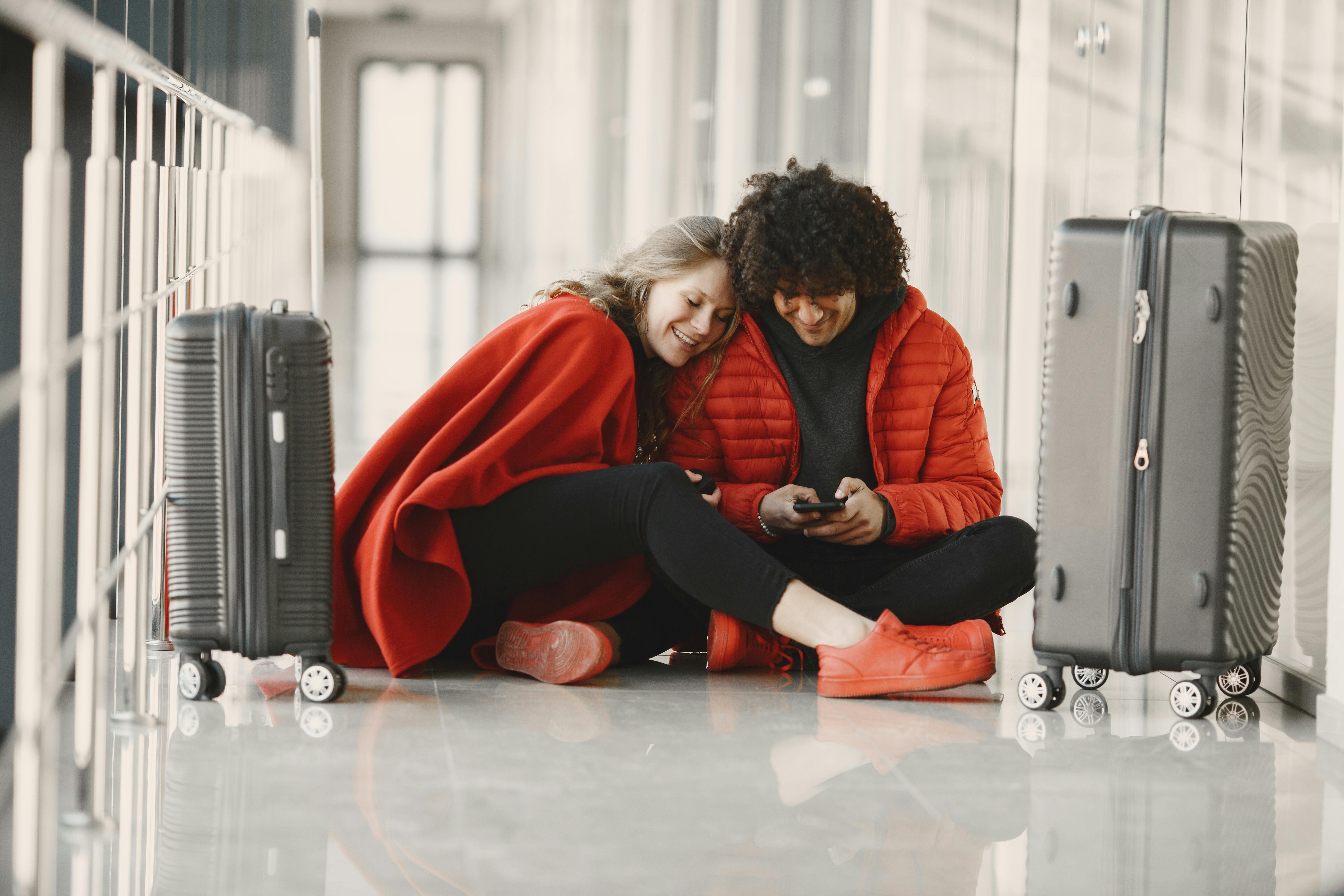 Happy couple sitting on the airport floor with luggage, sharing a moment using smartphone.