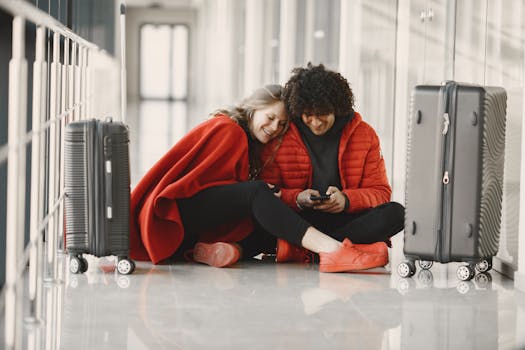 Happy couple sitting on the airport floor with luggage, sharing a moment using smartphone.