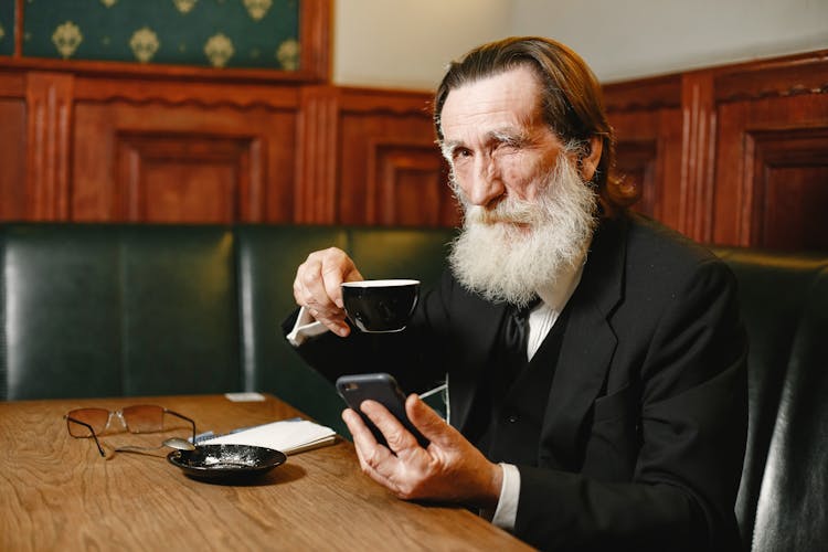 Man In Black Suit Holding Cellphone And A Cup While Winking On The Camera