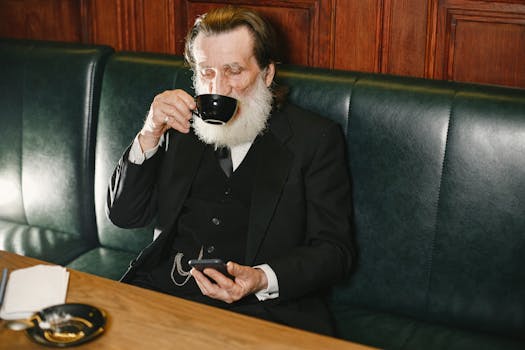 Senior man in a black suit enjoying coffee while sitting on a couch indoors.