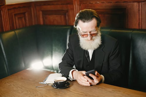 An Elderly Man in Black Suit Sitting on a Couch while Using Cellphone in front of a Wooden Table with Cup of Coffee