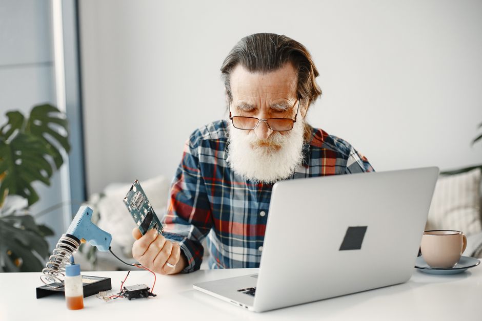 Elderly man with a beard works on a laptop, fixing electronic equipment at home.