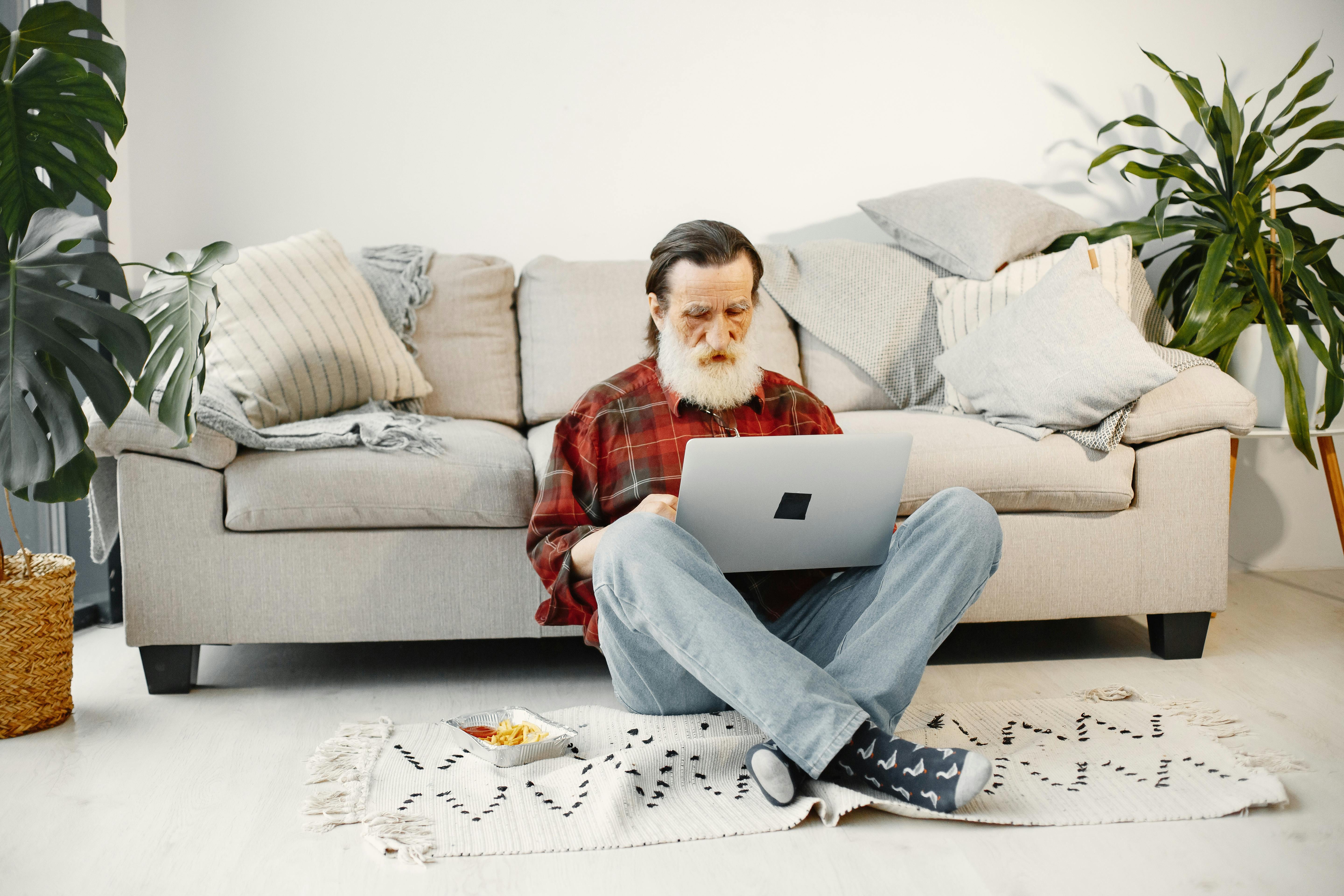 Senior man seated on floor with laptop, enjoying relaxed home environment with soft natural light.