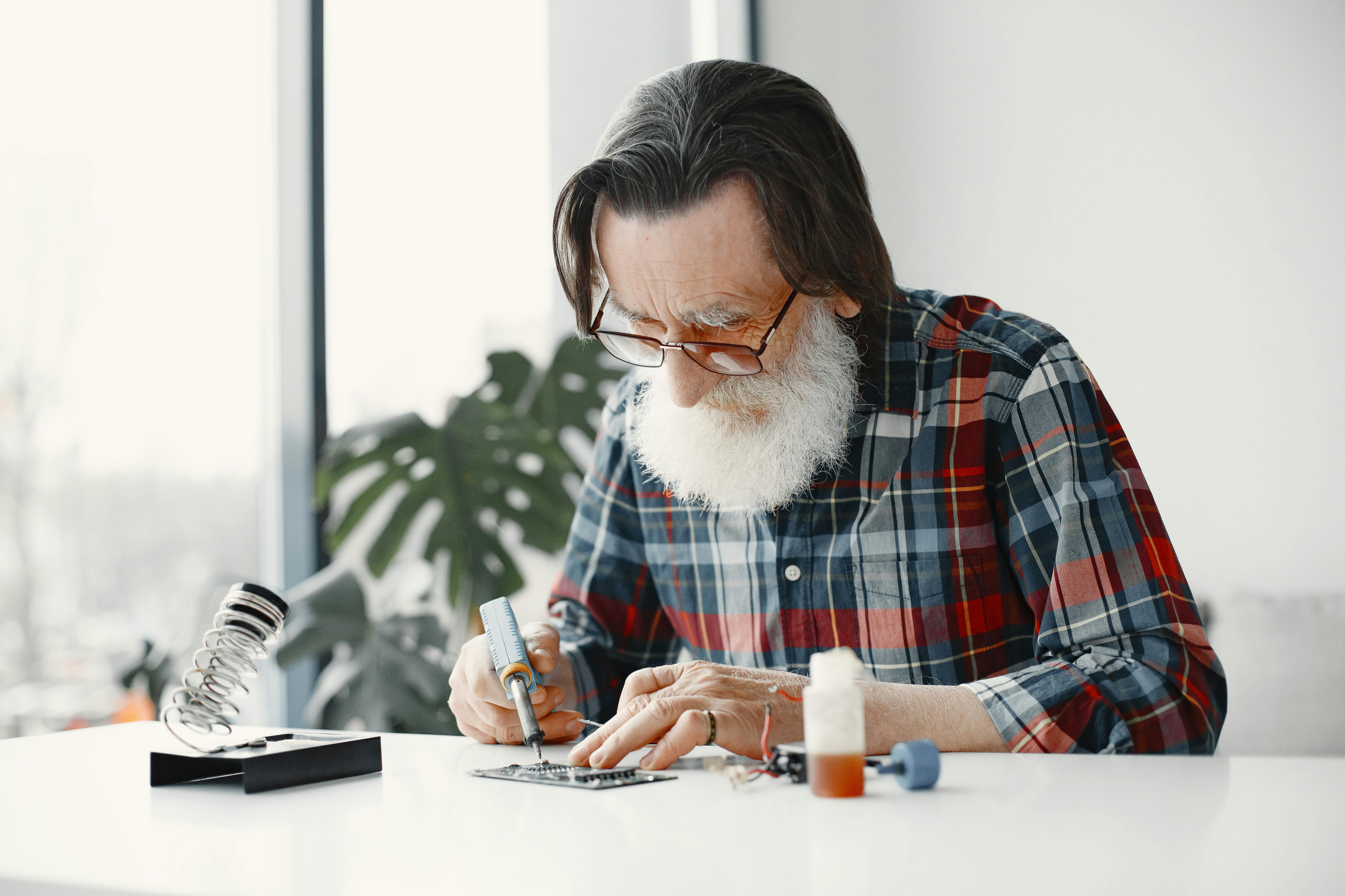 Elderly man fixing electronic device on table in bright room.