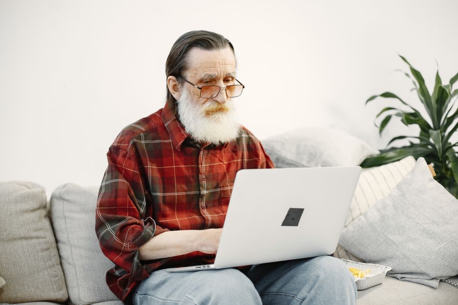 An elderly man using a laptop on a comfortable sofa in a home setting.