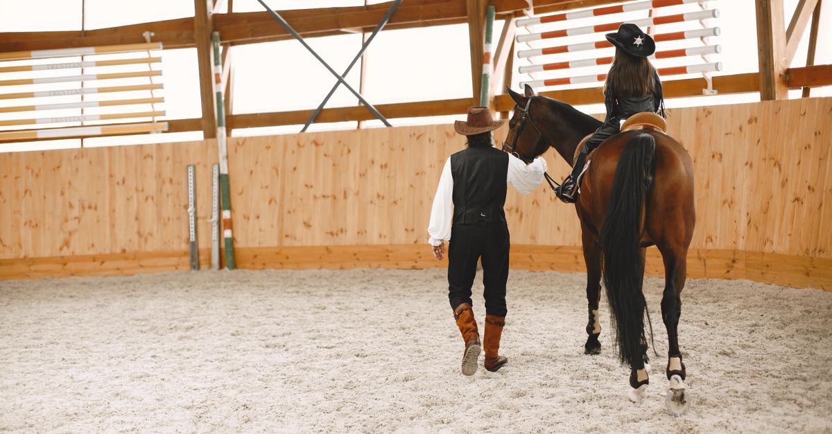 A girl riding a horse in an indoor arena with a man guiding them, both wearing cowboy hats.