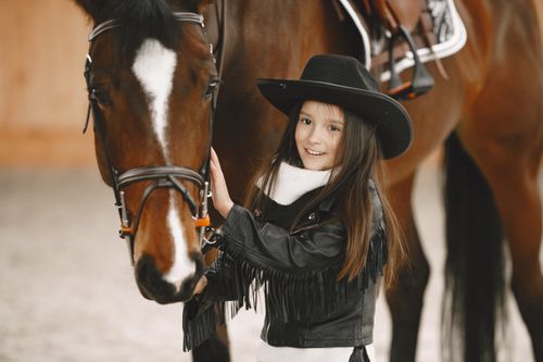 Free A cheerful young girl in a cowboy hat bonding with a horse indoors. Stock Photo