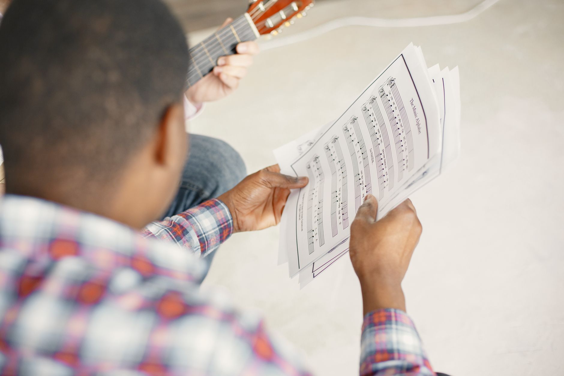 A close-up of a man holding sheet music while another person plays guitar indoors.