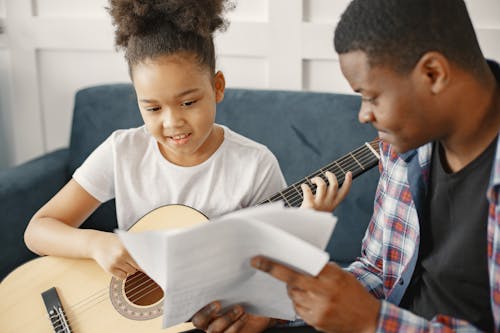 Girl Learning to Play Guitar Beside a Man Holding a Music Sheet