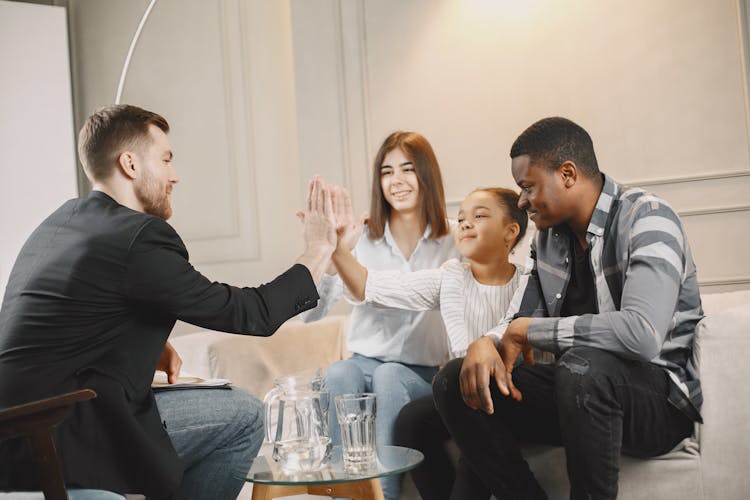 Little Girl Sitting With Parents On Sofa High-Fiving Guest