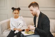 A child holding a plush toy listens to a man in a therapy session.