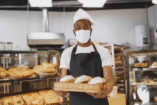 A baker wearing a face mask presenting fresh pastries in a modern bakery setting.