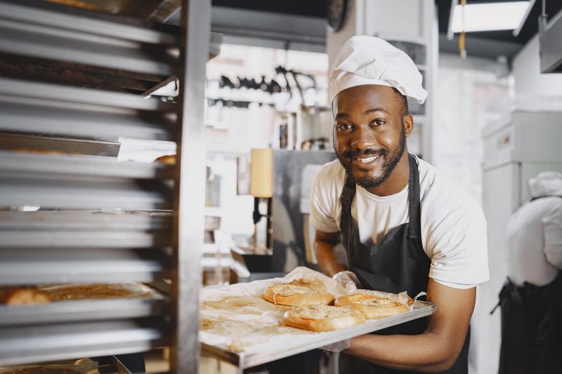 A cheerful baker presenting fresh breads from an industrial oven. Captured in a bakery setting.