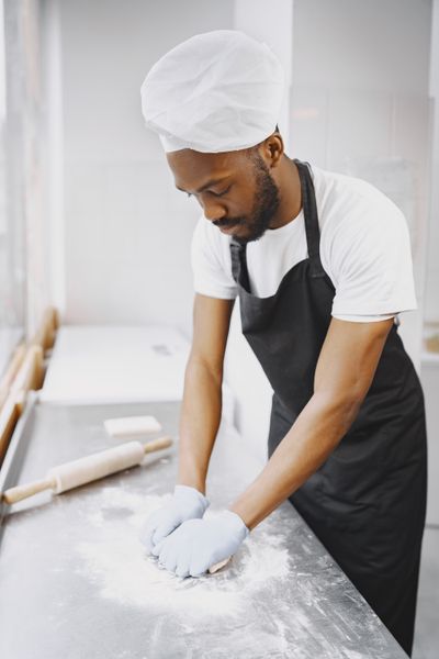 A baker in a white cap working dough on a bench in a bright bakery