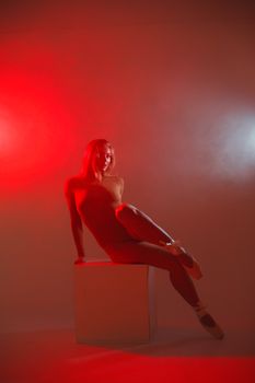Ballerina poses elegantly on a cube, illuminated by moody red lights, creating a striking visual contrast.