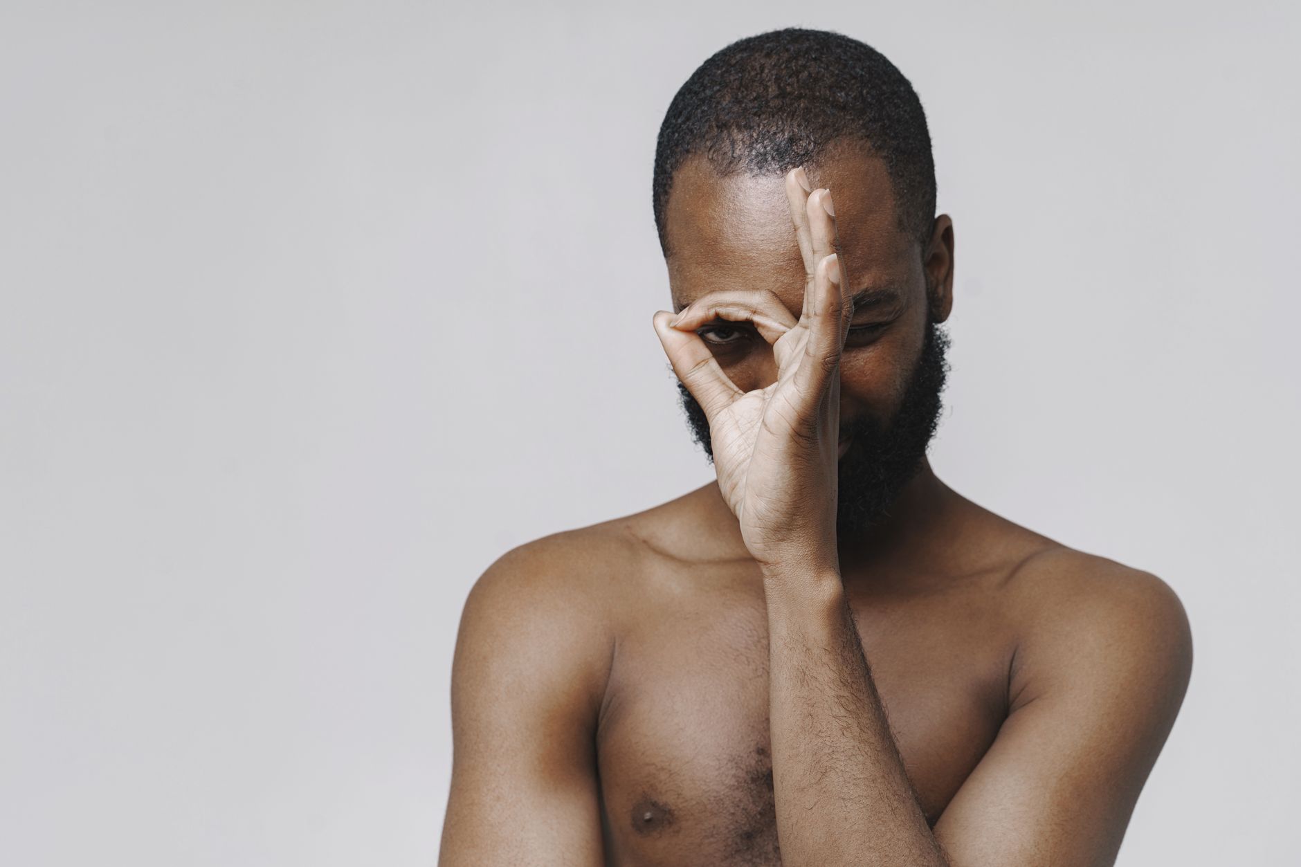 Studio portrait of a playful shirtless man making an OK sign over one eye.