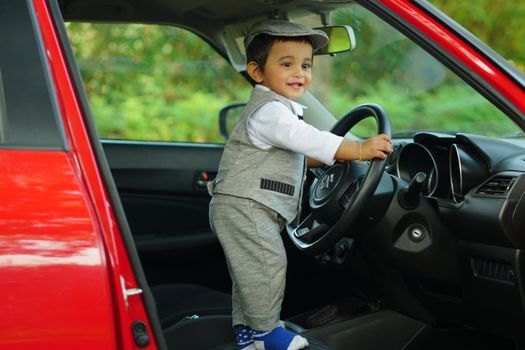 Cute child dressed in grey outfit playfully steering a red car outdoors.