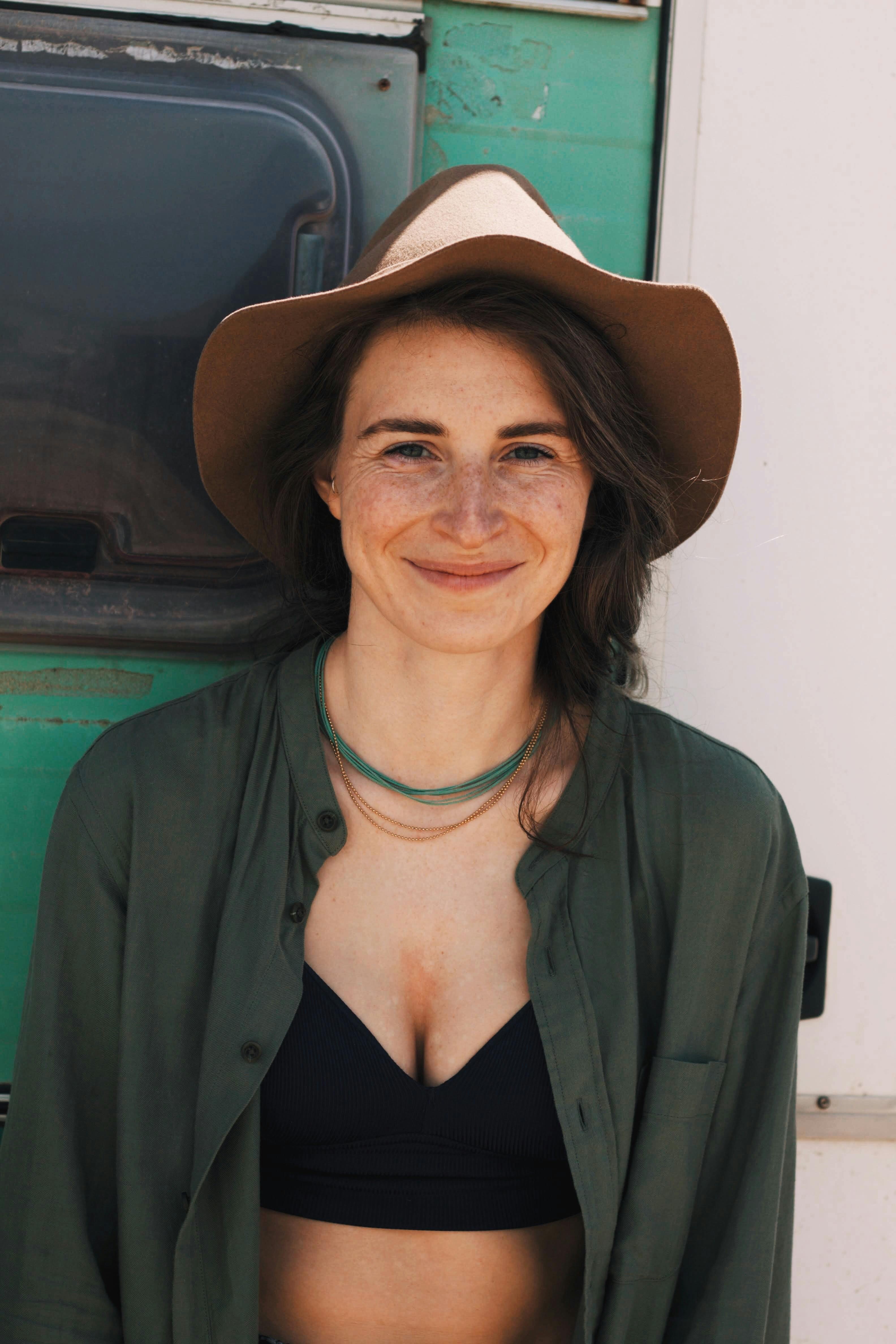 Positive young woman in trendy outfit and hat looking at camera while leaning on wall with window in daylight