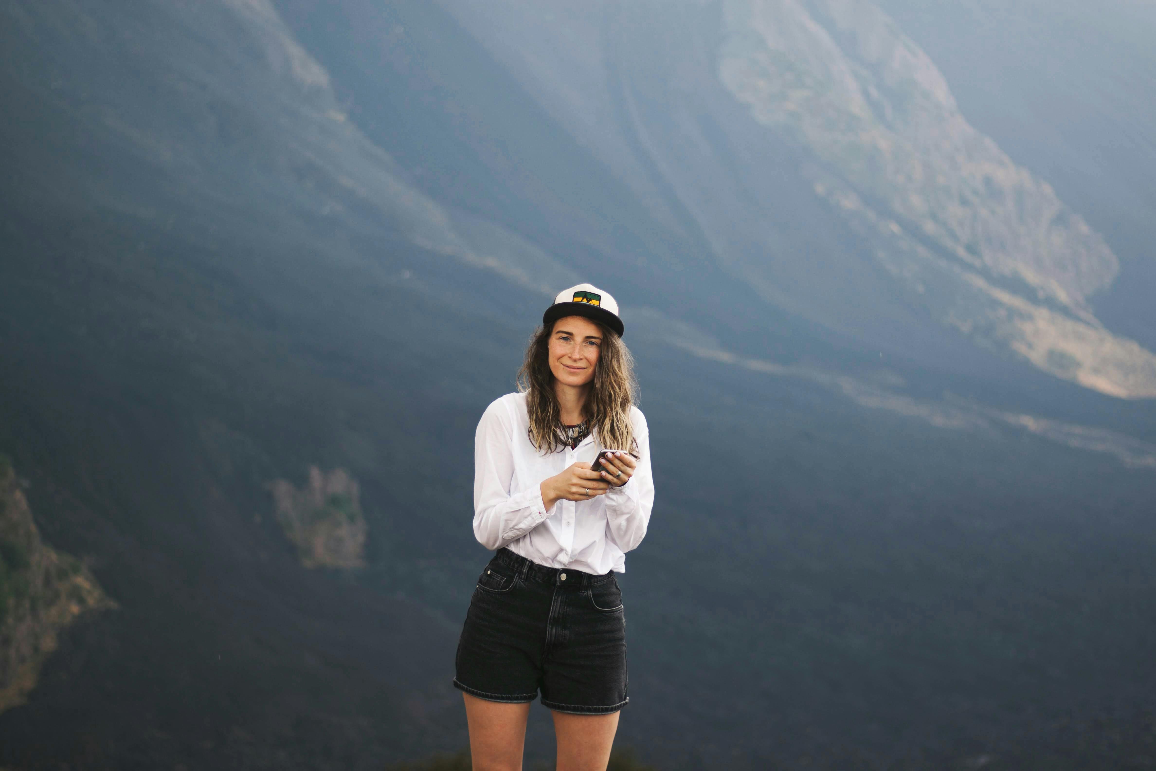 Woman in casual attire smiling while holding a phone in a mountainous landscape.