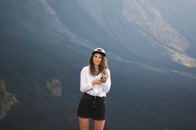 A Woman In White Long Sleeves And Black Shorts Smiling While Holding Her Phone