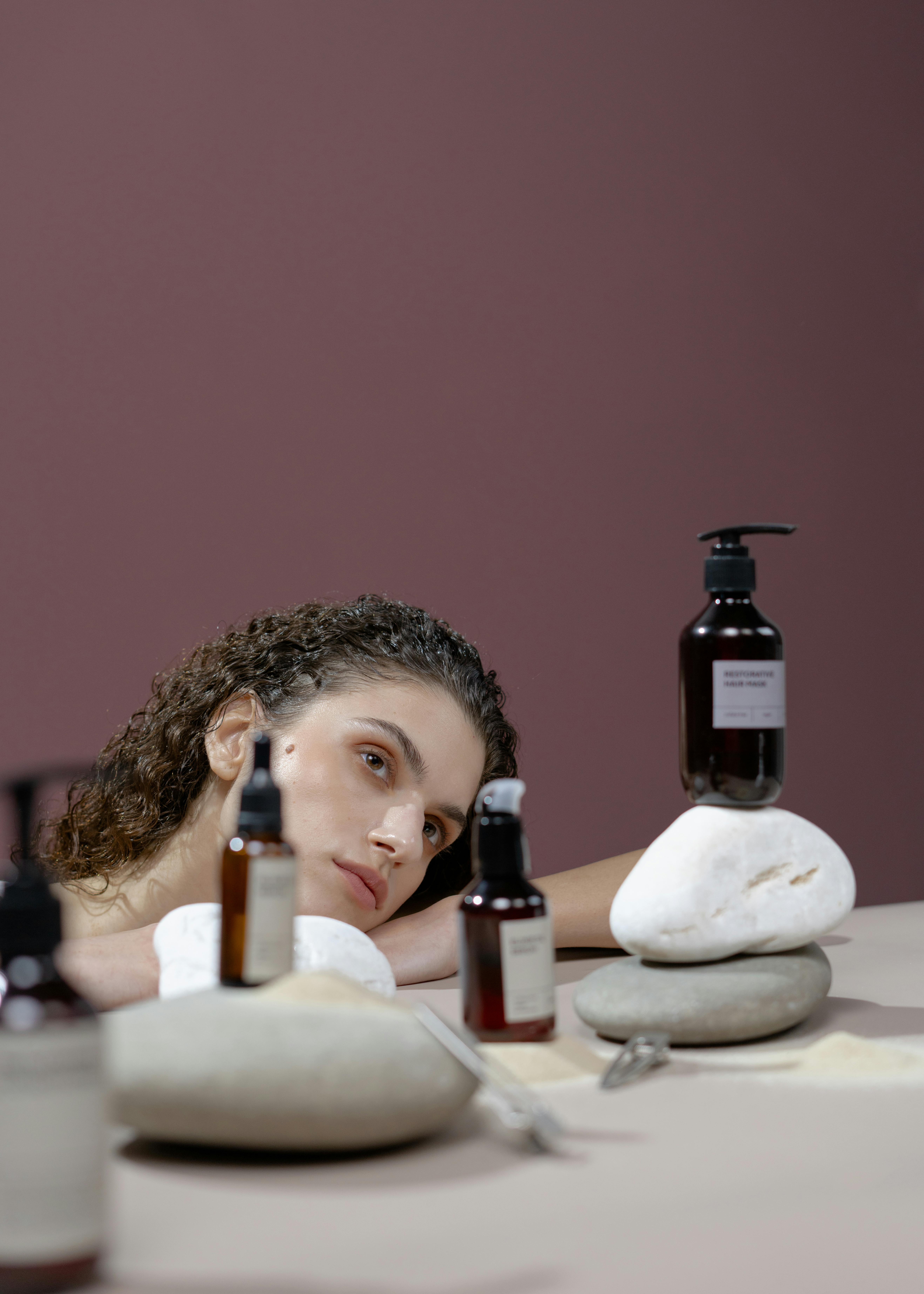 A woman with curly hair surrounded by skincare bottles on rocks, staring contemplatively.