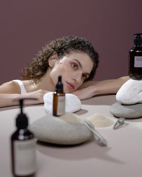 A woman with curly hair poses with skincare and hair products on a table.