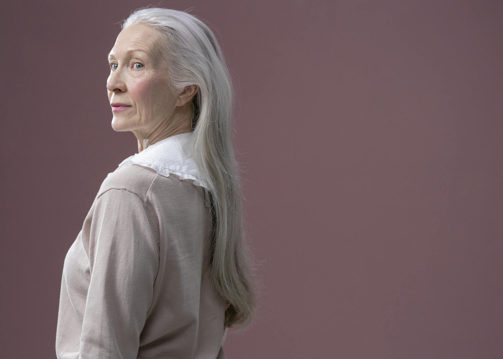 A sophisticated senior woman with long gray hair stands in profile against a solid background.