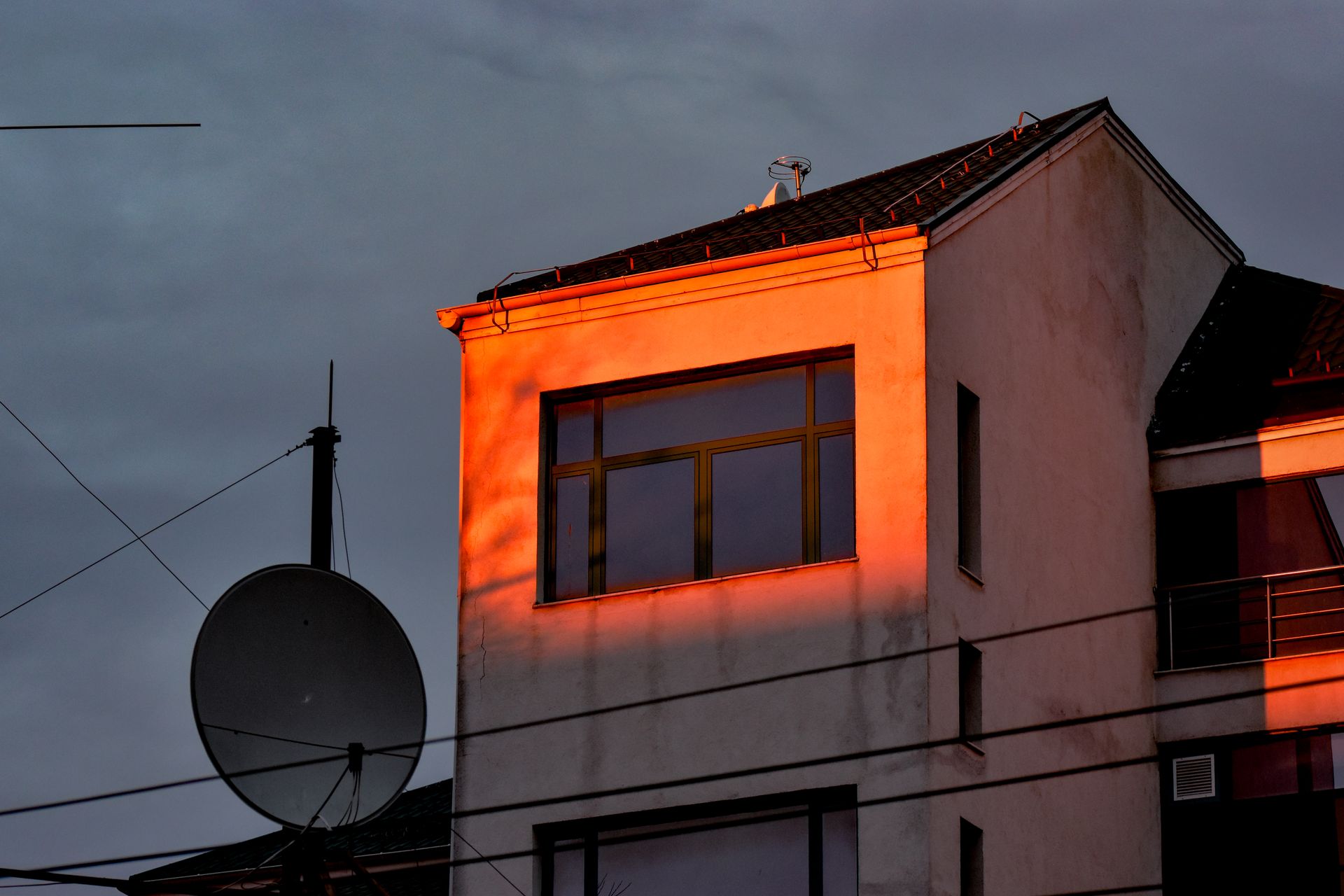 A modern building facade illuminated by sunset light with clouds and a satellite dish.
