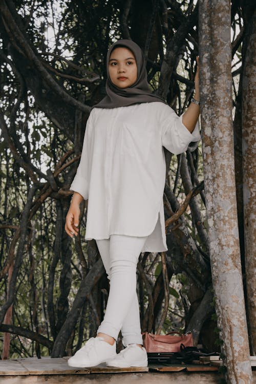 A Woman in White Clothes Wearing Hijab while Standing Near the Tree Trunk